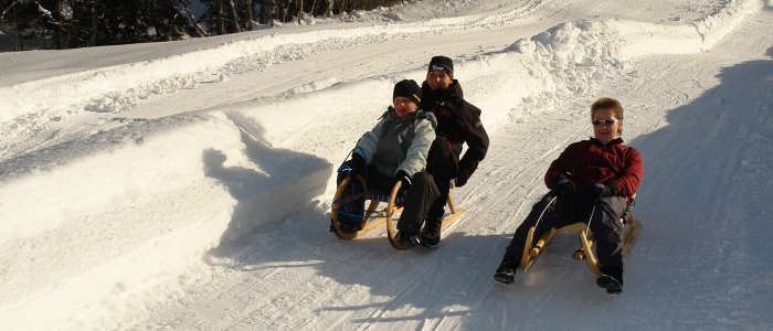 Drei Personen fahren an einem sonnigen Wintertag mit Holzschlitten eine schneebedeckte Bahn hinunter, umgeben von hohen Schneewänden.