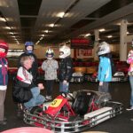 Eine Gruppe von Kindern mit Helmen und ein Mann versammeln sich um Gokarts in einer Indoor-Bahn; ein Kind steht neben einem Gokart mit der Nummer 3.