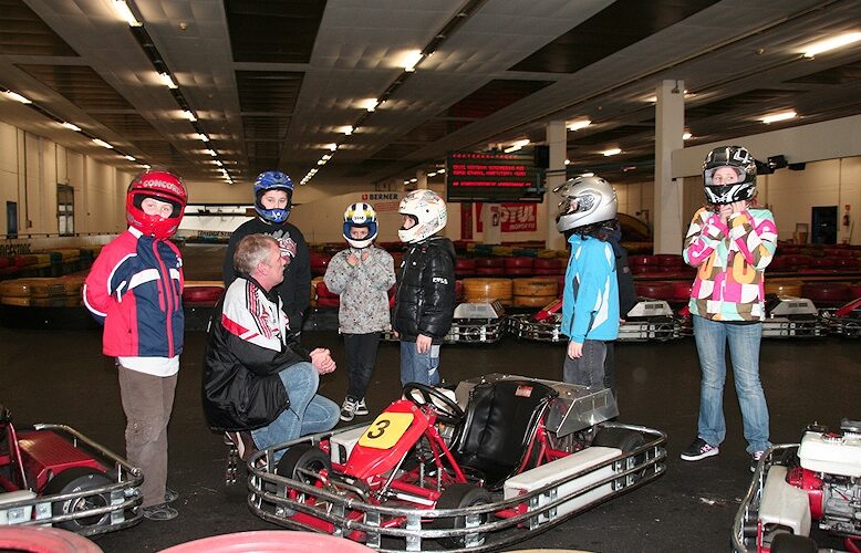 Eine Gruppe von Kindern mit Helmen und ein Mann versammeln sich um Gokarts in einer Indoor-Bahn; ein Kind steht neben einem Gokart mit der Nummer 3.
