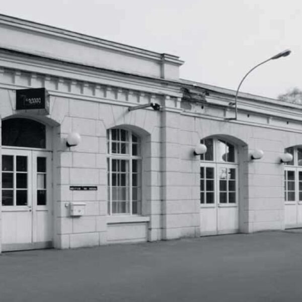 Ein weißes, einstöckiges Gebäude mit gewölbten Fenstern und Türen, Wandleuchten und einer Straßenlaterne davor. Der Himmel scheint bedeckt zu sein.