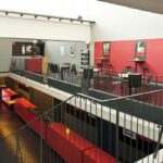 Modernes Zwischengeschoss im Innenbereich mit roten und grauen Wänden, schwarzem Geländer, hohen Tischen mit Hockern und einer Treppe, die in eine untere Etage führt. An den Wänden sind Plakate angebracht.