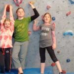 Drei Kinder stehen auf einer blauen Matte vor einer Indoor-Kletterwand, halten sich an den Händen und heben die Arme.