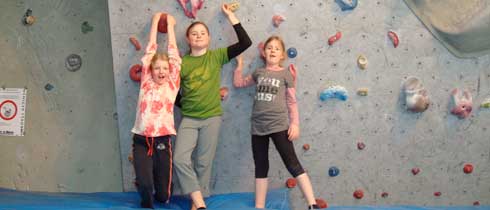 Drei Kinder stehen auf einer blauen Matte vor einer Indoor-Kletterwand, halten sich an den Händen und heben die Arme.