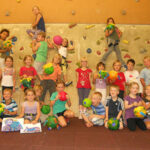Eine Gruppe Kinder posiert drinnen vor einer Kletterwand, hält bunte Bastelarbeiten in den Händen und lächelt in die Kamera.