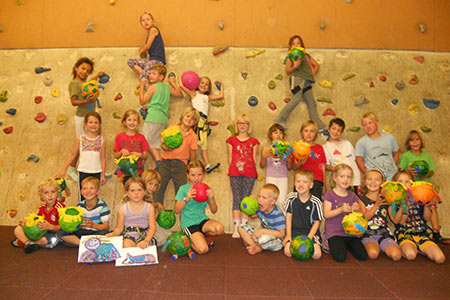 Eine Gruppe Kinder posiert drinnen vor einer Kletterwand, hält bunte Bastelarbeiten in den Händen und lächelt in die Kamera.