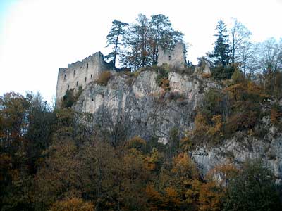 Die Ruinen einer steinernen Burg liegen auf einer felsigen Klippe, umgeben von Herbstbäumen mit Blättern in Braun-, Gelb- und Grüntönen.