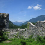 Im Vordergrund stehen steinerne Ruinen, im Hintergrund sind Berge und ein blauer Himmel zu sehen. Grüne Vegetation umgibt das Bauwerk.
