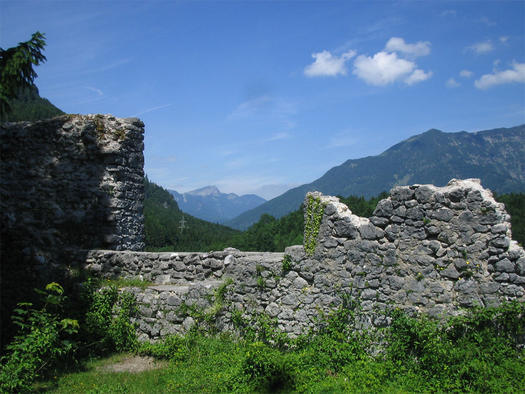 Im Vordergrund stehen steinerne Ruinen, im Hintergrund sind Berge und ein blauer Himmel zu sehen. Grüne Vegetation umgibt das Bauwerk.