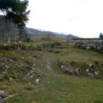 Grasbedeckte Ruinen mit Steinmauern und Stufen, im Freien gelegen, mit einigen Bergen im Hintergrund unter einem bewölkten Himmel.