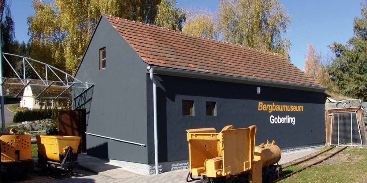 Ein dunkelgraues Gebäude mit der Aufschrift "Bergbaumuseum Goberling" steht neben Bergbaugeräten und Bahngleisen, umgeben von Bäumen mit Herbstlaub.