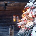 Ein schneebedeckter, mit roten Ornamenten und Kerzen geschmückter Weihnachtsbaum steht vor einer rustikalen Holzhütte mit einem beleuchteten Fenster und einem Schlitten in der Nähe.