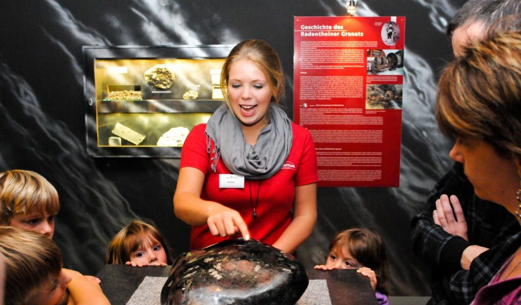 Ein Museumsführer im roten Hemd erklärt einer Gruppe von Kindern und Erwachsenen ein großes Gesteins- oder Mineralienexemplar, während im Hintergrund eine Informationsanzeige zu sehen ist.