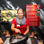 Ein Museumsführer im roten Hemd erklärt einer Gruppe von Kindern und Erwachsenen ein großes Gesteins- oder Mineralienexemplar, während im Hintergrund eine Informationsanzeige zu sehen ist.