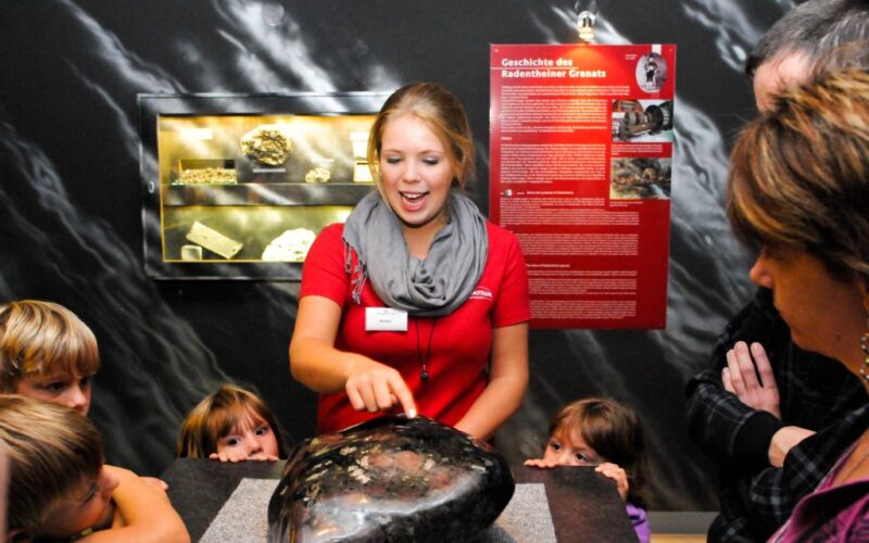 Ein Museumsführer im roten Hemd erklärt einer Gruppe von Kindern und Erwachsenen ein großes Gesteins- oder Mineralienexemplar, während im Hintergrund eine Informationsanzeige zu sehen ist.