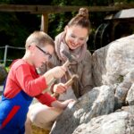 Eine Frau und ein Kind, die Schutzbrillen tragen, benutzen kleine Hämmer, um an einem sonnigen Tag im Freien große Felsen abzuschlagen.