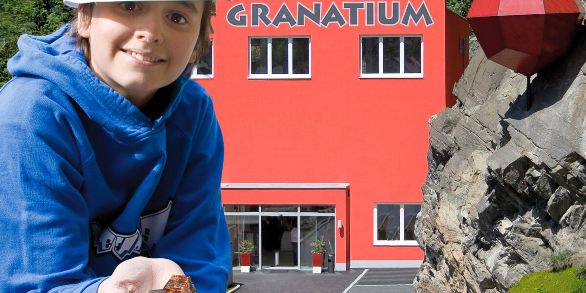 Ein Kind, das einen weißen Schutzhelm trägt, hält Steine vor einem leuchtend roten Gebäude mit der Aufschrift "Granatium", neben dem ein großes rotes geometrisches Gebilde an einem felsigen Abhang befestigt ist.