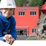 Ein Kind, das einen weißen Schutzhelm trägt, hält Steine vor einem leuchtend roten Gebäude mit der Aufschrift "Granatium", neben dem ein großes rotes geometrisches Gebilde an einem felsigen Abhang befestigt ist.