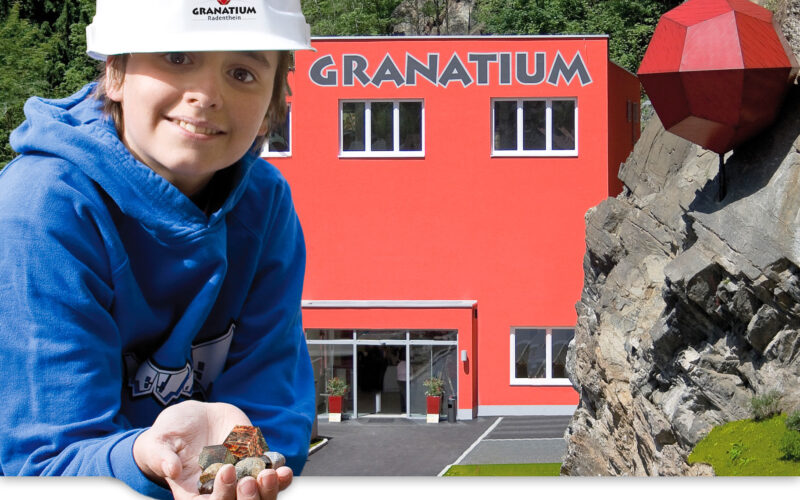 Ein Kind, das einen weißen Schutzhelm trägt, hält Steine vor einem leuchtend roten Gebäude mit der Aufschrift "Granatium", neben dem ein großes rotes geometrisches Gebilde an einem felsigen Abhang befestigt ist.