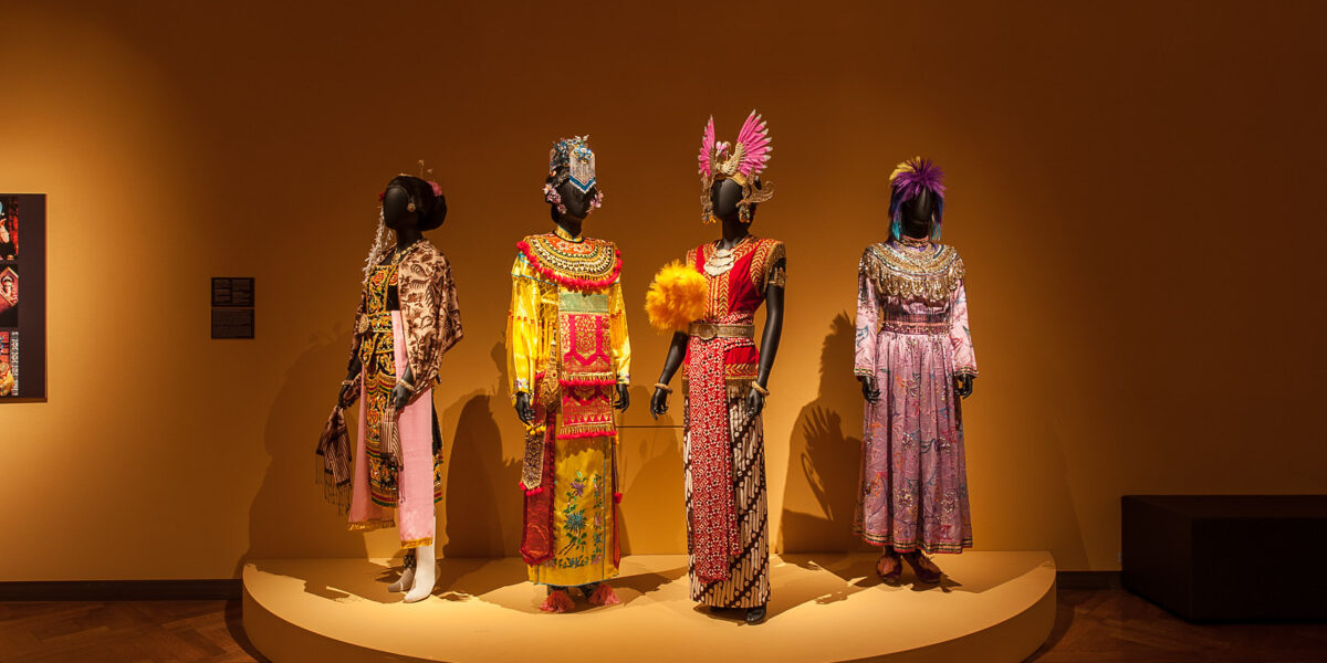 Vier Schaufensterpuppen tragen kunstvolle, farbenfrohe Trachten mit aufwendigen Kopfbedeckungen und stehen auf einem runden Podest vor einer schlichten braunen Wand in einer Museumsumgebung.
