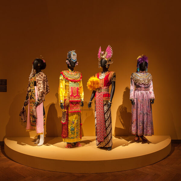 Vier Schaufensterpuppen tragen kunstvolle, farbenfrohe Trachten mit aufwendigen Kopfbedeckungen und stehen auf einem runden Podest vor einer schlichten braunen Wand in einer Museumsumgebung.