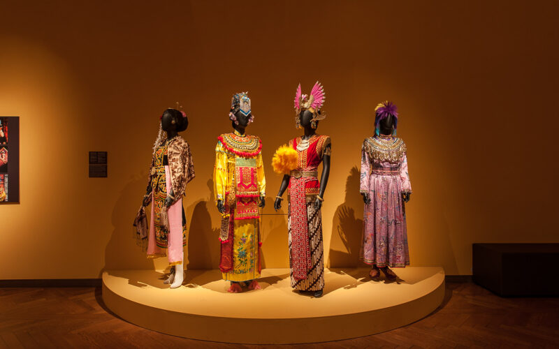 Vier Schaufensterpuppen tragen kunstvolle, farbenfrohe Trachten mit aufwendigen Kopfbedeckungen und stehen auf einem runden Podest vor einer schlichten braunen Wand in einer Museumsumgebung.