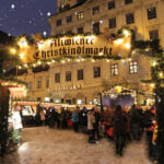 Menschenmengen versammeln sich unter dem beleuchteten Schild "Altwiener Christkindlmarkt" auf einem verschneiten Weihnachtsmarkt im Freien vor einem historischen Gebäude in der Abenddämmerung.