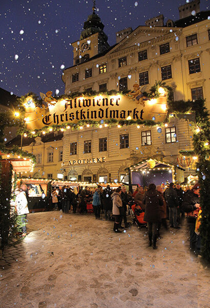 Menschenmengen versammeln sich unter dem beleuchteten Schild "Altwiener Christkindlmarkt" auf einem verschneiten Weihnachtsmarkt im Freien vor einem historischen Gebäude in der Abenddämmerung.