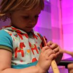 Ein kleines Kind in einem bunten Hemd spielt eine Holzblockflöte, während eine Erwachsenenhand auf das Instrument zeigt. Der Hintergrund ist mit rosa und lila Lichtern beleuchtet.