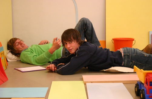 In einem bunten Raum liegen zwei Jugendliche auf dem Boden, einer auf dem Rücken, der andere auf dem Bauch. In der Nähe befinden sich Papiere, ein Klemmbrett und ein Spielzeug-LKW.