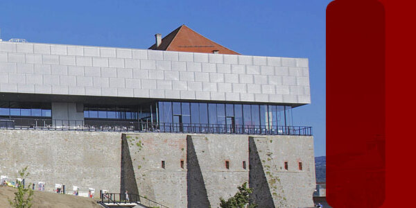 Ein modernes Gebäude mit einer Fassade aus Glas und Metall thront auf einer alten Steinmauer unter einem klaren blauen Himmel. Zwei große rote Rechtecke überlagern die rechte Seite des Bildes.