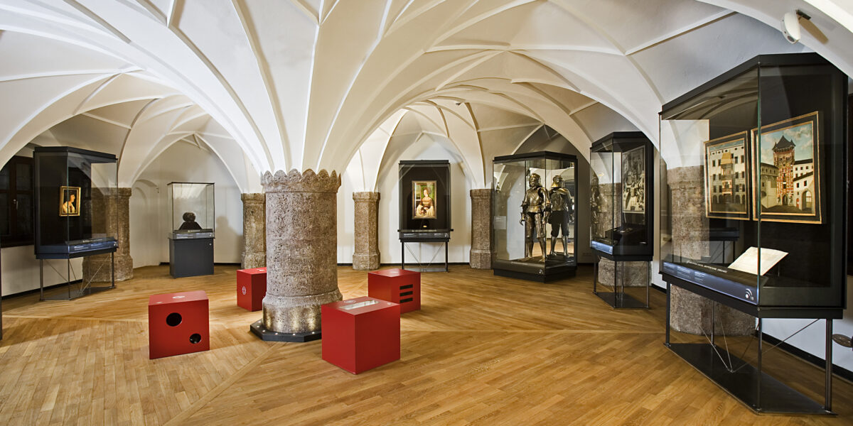 Museumsgalerie mit Gewölbedecken, Rüstungsauslage, gerahmten Kunstwerken, Büstenskulpturen und Glasvitrinen auf einem Holzboden. Rote Sitzwürfel sind im ganzen Raum verteilt.