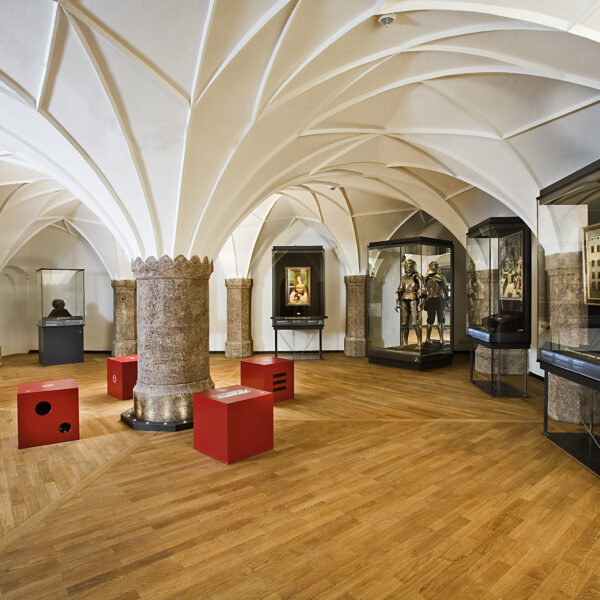 Museumsgalerie mit Gewölbedecken, Rüstungsauslage, gerahmten Kunstwerken, Büstenskulpturen und Glasvitrinen auf einem Holzboden. Rote Sitzwürfel sind im ganzen Raum verteilt.