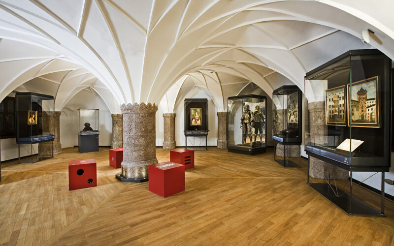 Museumsgalerie mit Gewölbedecken, Rüstungsauslage, gerahmten Kunstwerken, Büstenskulpturen und Glasvitrinen auf einem Holzboden. Rote Sitzwürfel sind im ganzen Raum verteilt.