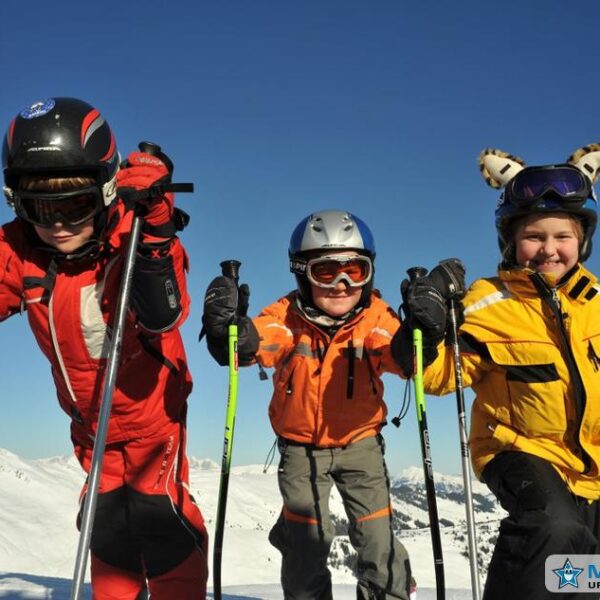 Drei Kinder in Skikleidung posieren mit Skistöcken auf einem verschneiten Berghang unter strahlend blauem Himmel.