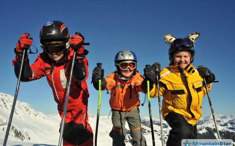 Drei Kinder in Skikleidung posieren mit Skistöcken auf einem verschneiten Berghang unter strahlend blauem Himmel.