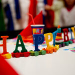 Bunte Buchstaben mit der Aufschrift „HAPPY“ und dekorierte Becher mit Strohhalmen sind auf einem Tisch angeordnet und vermitteln den Eindruck einer festlichen Geburtstagsparty.