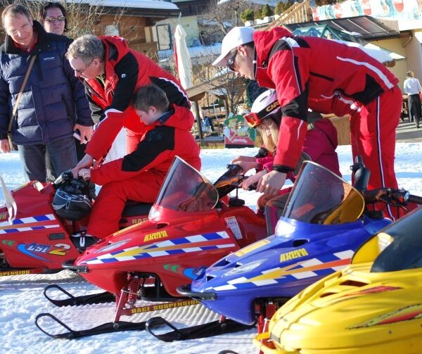 Zwei Erwachsene helfen Kindern, sich in einem verschneiten Außenbereich auf kleine Motorschlitten zu setzen, während ein anderer Erwachsener in der Nähe beobachtet.