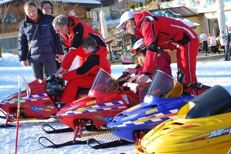 Zwei Erwachsene helfen Kindern, sich in einem verschneiten Außenbereich auf kleine Motorschlitten zu setzen, während ein anderer Erwachsener in der Nähe beobachtet.