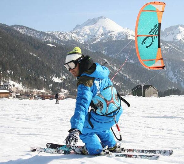 Eine Person in blauer Winterkleidung fährt auf Skiern durch eine verschneite Landschaft mit Bergen und Gebäuden im Hintergrund.