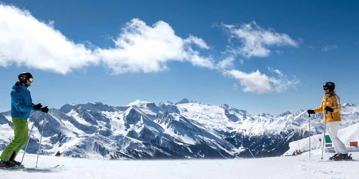 Zwei Skifahrer stehen sich auf einer verschneiten Piste mit schneebedeckten Bergen und einem blauen Himmel im Hintergrund gegenüber.