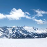 Zwei Skifahrer stehen sich auf einer verschneiten Piste mit schneebedeckten Bergen und einem blauen Himmel im Hintergrund gegenüber.