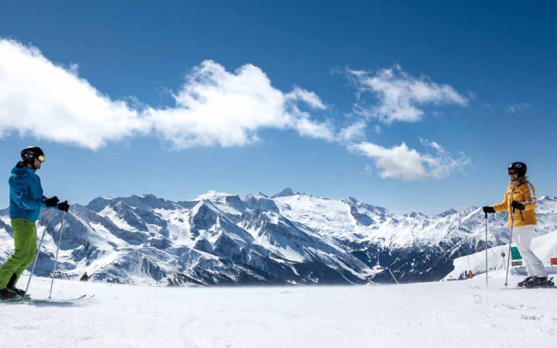 Zwei Skifahrer stehen sich auf einer verschneiten Piste mit schneebedeckten Bergen und einem blauen Himmel im Hintergrund gegenüber.