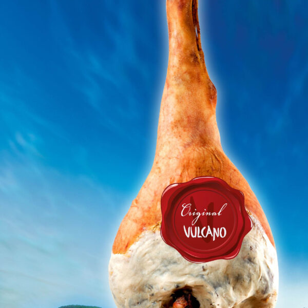 Ein ganzer Rohschinken mit einem roten "Original Vulcano"-Etikett hängt vor einem blauen Himmel und einem grünen Landschaftshintergrund.