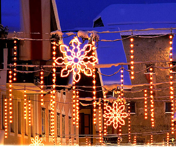 Dekorative orangefarbene Lichter und beleuchtete Schneeflocken hängen in der Abenddämmerung über einer verschneiten, von Gebäuden gesäumten Straße.