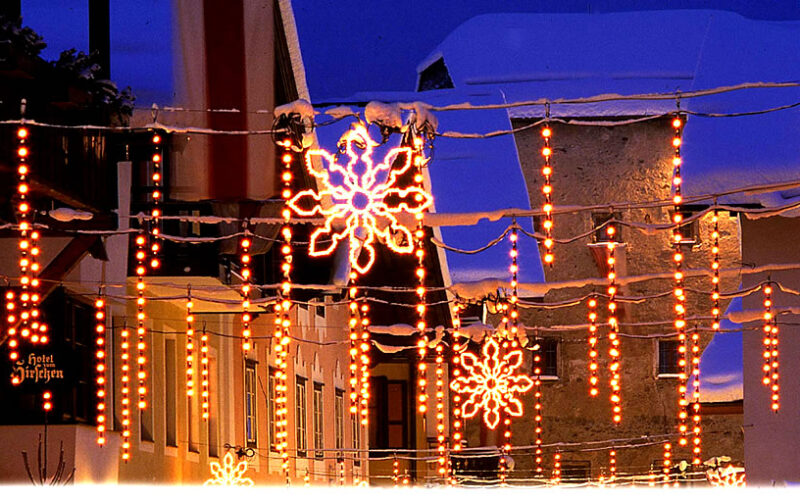 Dekorative orangefarbene Lichter und beleuchtete Schneeflocken hängen in der Abenddämmerung über einer verschneiten, von Gebäuden gesäumten Straße.