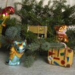 Ein mit Ornamenten verzierter Tannenzweig, darunter ein Papagei, ein Radio, ein Bär, eine Katze und ein Koffer, ruht auf einer grauen Fläche.