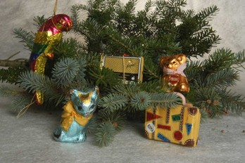 Ein mit Ornamenten verzierter Tannenzweig, darunter ein Papagei, ein Radio, ein Bär, eine Katze und ein Koffer, ruht auf einer grauen Fläche.