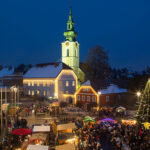 Eine nächtliche Szene eines Weihnachtsmarktes auf einem Stadtplatz mit einer Menschenmenge, geschmückten Ständen, einem beleuchteten Weihnachtsbaum und einer Kirche mit beleuchtetem Uhrenturm im Hintergrund.