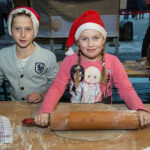 Zwei Kinder mit festlichen Weihnachtsmannmützen stehen an einem Tisch, wobei ein Mädchen mit einem Nudelholz Teig ausrollt, während der Junge zusieht.