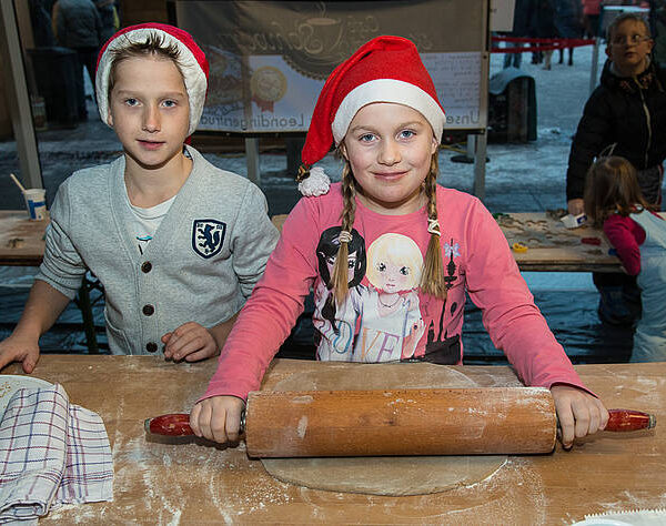 Zwei Kinder mit festlichen Weihnachtsmannmützen stehen an einem Tisch, wobei ein Mädchen mit einem Nudelholz Teig ausrollt, während der Junge zusieht.